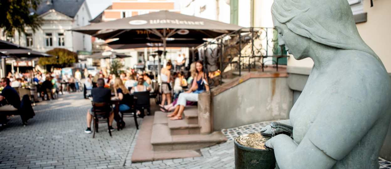 Das Bild zeigt die Statue einer Marktfrau. Sie hält einen Krug mit Wasser. Im Hintergrund sitzen einige Menschen in den Biergärten der Gastwirtschaften am Markptlatz.  