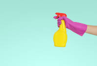 Bild mit hellblaum Hintergrund. Eine Hand im lila Handschuh hält eine gelbe Sprühflasche